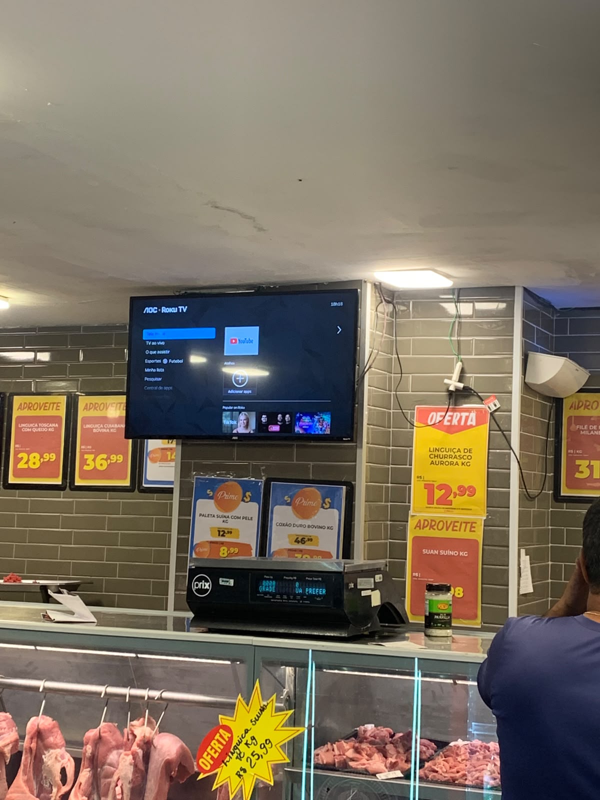 TV num açougue mostrando a tela inicial do Roku, parada, cercada de cartazes de oferta de carne.